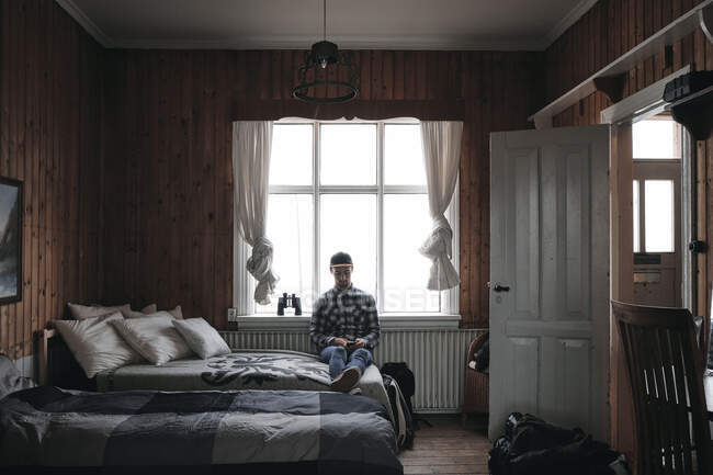 Hombre adulto sentado y descansando en la cama mientras usa el teléfono inteligente en casa en Islandia - foto de stock