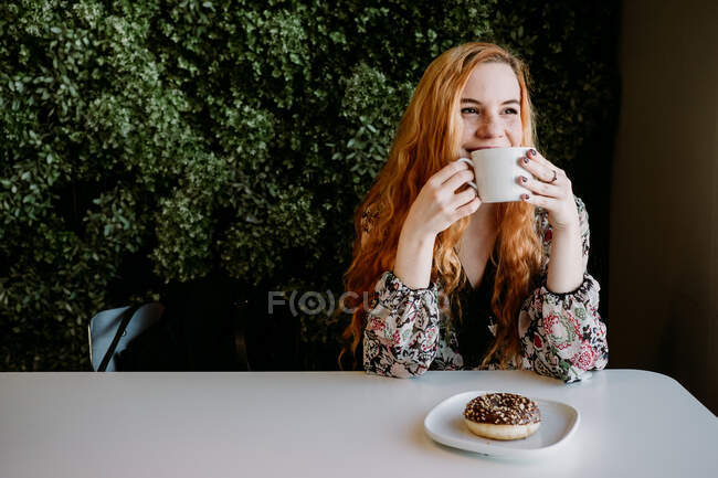 Jolie rousse gaie jolie femme avec tasse assise à la brousse et regardant loin. — Photo de stock