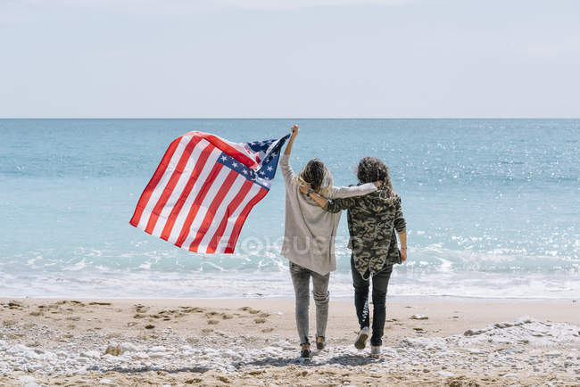 Vista trasera de mujeres jóvenes caminando en la playa con bandera de EE.UU. . - foto de stock