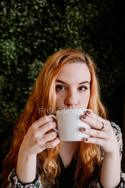 Fröhliche rothaarige hübsche Frau mit Tasse sitzt am Busch und schaut weg. — Stockfoto
