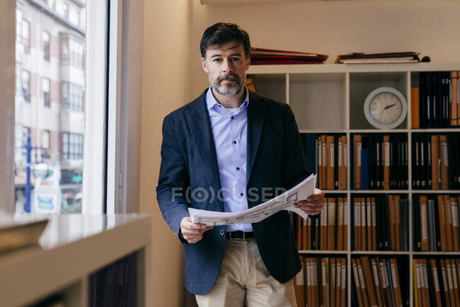 Hombre adulto guapo de pie en la oficina con documentos y mirando a la cámara. - foto de stock