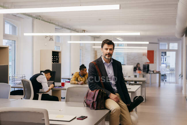 Красивый бородатый взрослый мужчина сидит на столе в офисе и смотрит в камеру. — стоковое фото