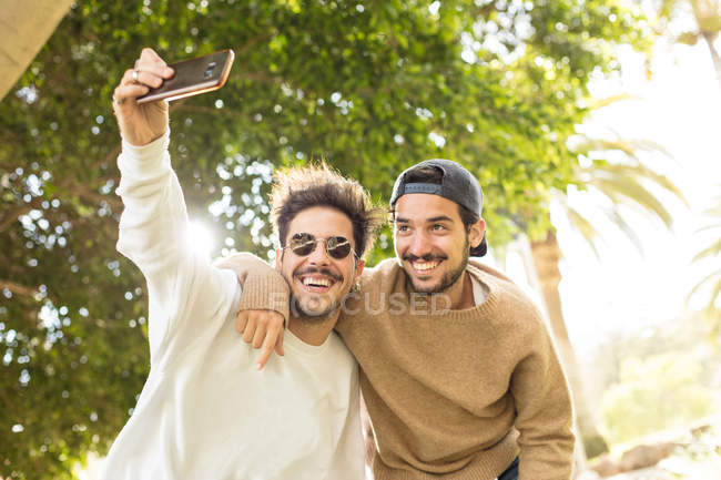 Sorridenti amici maschi felici scattare selfie con smartphone nel parco soleggiato — Foto stock