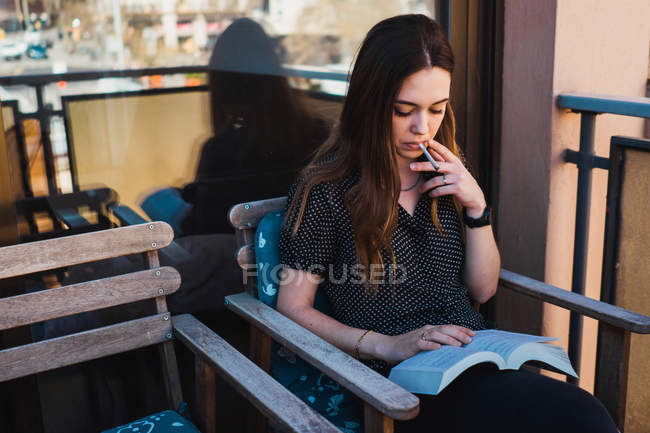 Жінка читає книгу і курить на дерев'яному стільці на балконі — стокове фото