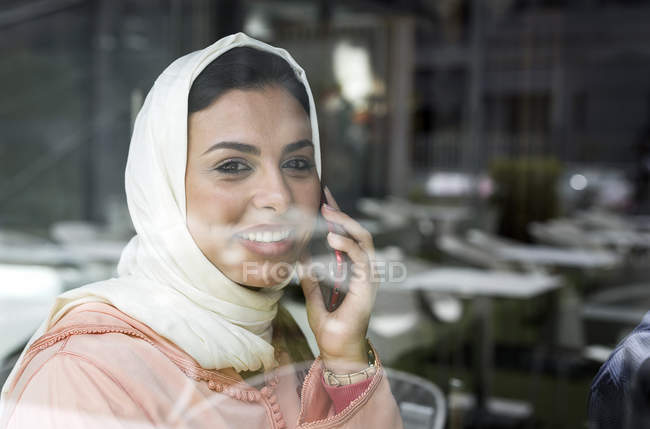 Марокканская женщина с хиджабом и традиционным арабским платьем разговаривает по телефону за стеклом — стоковое фото