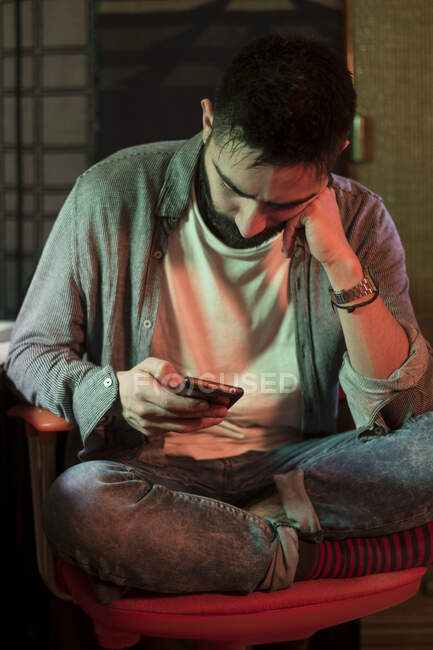 Вдумчивый взрослый мужчина сидит в кресле и просматривает смартфон в помещении. — стоковое фото