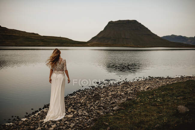 Вид сзади женщины в белом платье, стоящей на спокойном озере на склоне холма. — стоковое фото