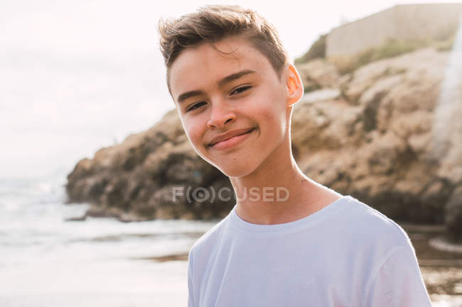 Retrato de niño lindo sonriente en camiseta blanca de pie en la orilla del mar en verano - foto de stock