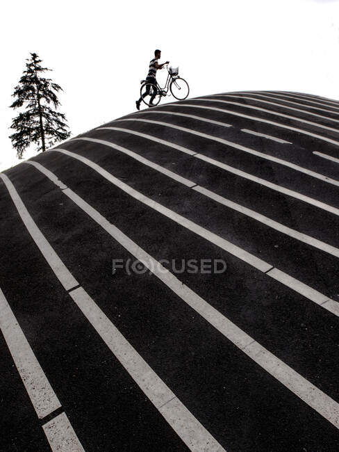 Black and white concept photo of man walking with bicycle up on hill in road pavement with white marking lines — Stock Photo