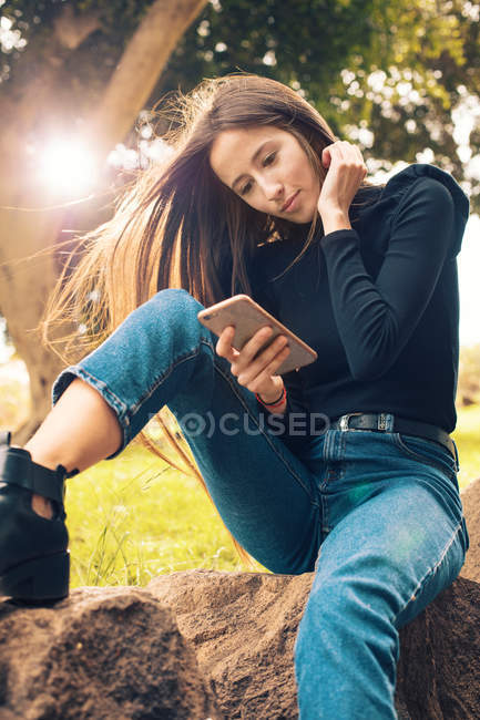 Mujer joven sentada en la roca y usando el teléfono inteligente en el parque - foto de stock