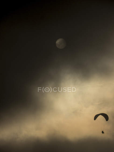 Petite silhouette noire de personne parapente avec parachute dans les nuages sombres avec cercle blanc de lune — Photo de stock