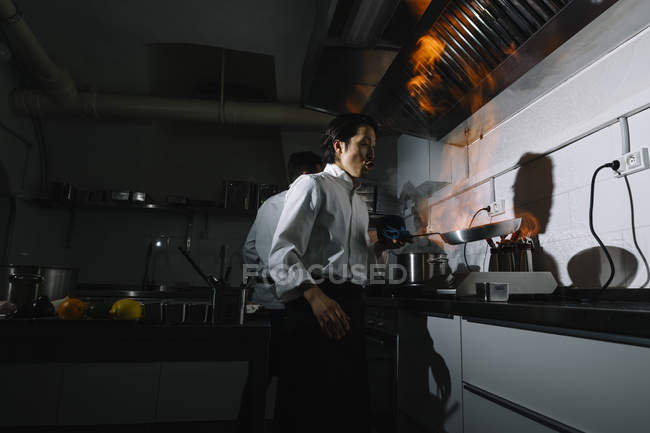 Cook making a flambe in restaurant kitchen with colleague on background — Stock Photo