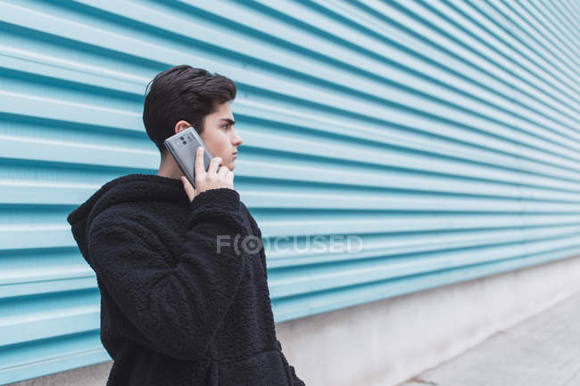 Молодой подросток, стоящий у металлической стены и разговаривающий на смартфоне на улице — стоковое фото