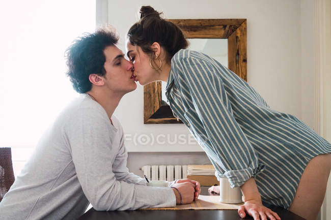Joven hombre y mujer besándose en la mesa con tazas - foto de stock