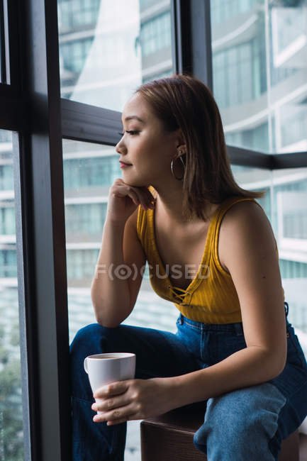 Вдумчивая молодая женщина с чашкой сидя в квартире и глядя в окно — стоковое фото