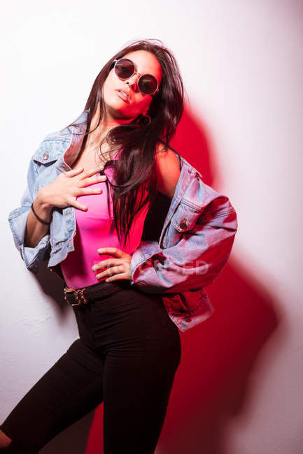 Pretty young pink dressed woman in sunglasses standing at white wall and looking at camera — Stock Photo