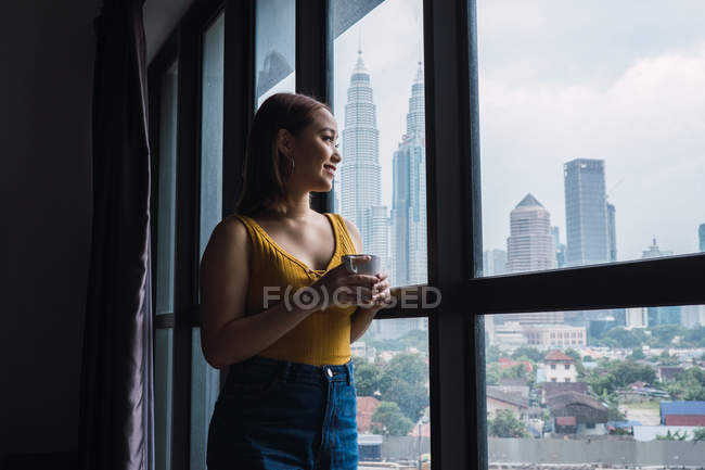 Усміхаючись досить Азіатський жінка, стоячи з Кубок на вікна і дивитися вбік — стокове фото