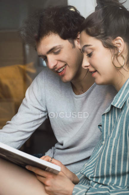 Feliz joven hombre y mujer sentados juntos y leyendo el libro en casa - foto de stock