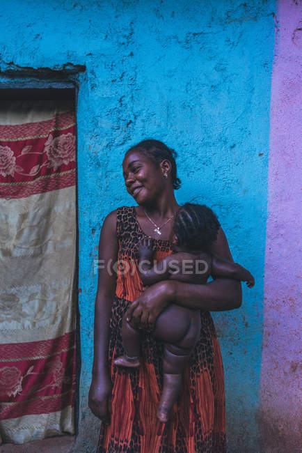 CAMARÕES - ÁFRICA - ABRIL 5, 2018: Mulher africana adulta sorridente segurando criança nua e em pé na parede colorida na rua — Fotografia de Stock