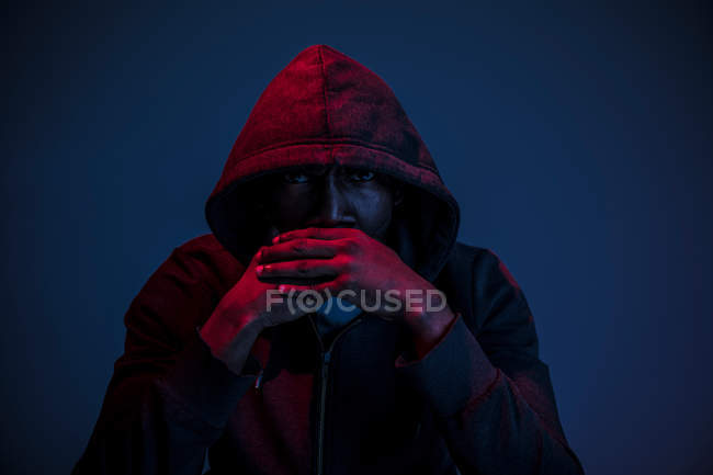 Retrato del hombre de confianza en la capucha con las manos apiladas sentado sobre fondo oscuro - foto de stock