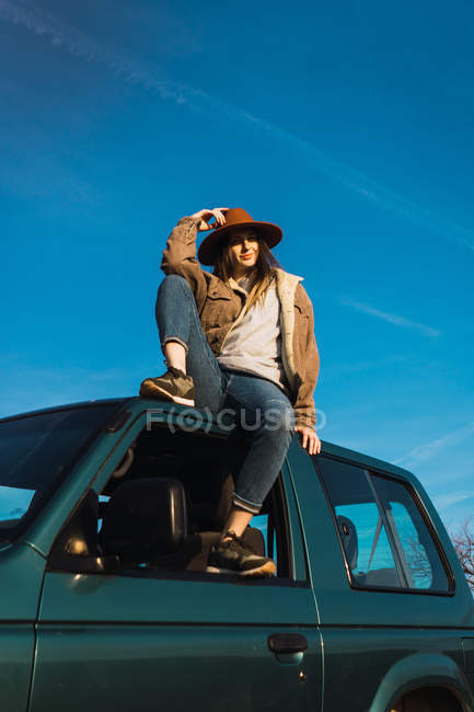 Stylish woman in hat sitting on car roof — Stock Photo