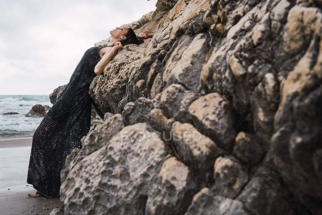Femme à la mode en robe noire appuyée sur le rocher sur la plage — Photo de stock