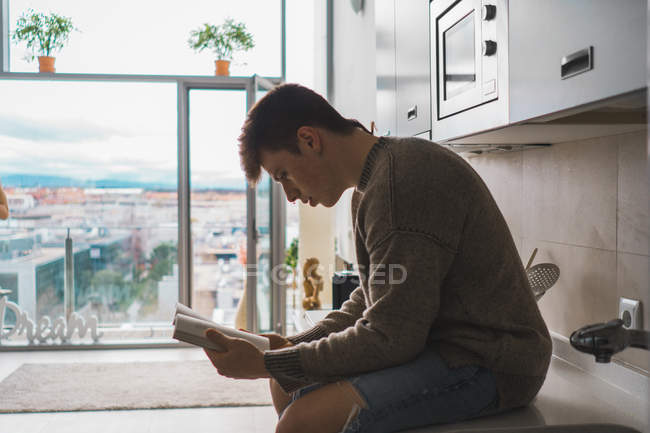 Young man in sweater and jeans sitting on kitchen counter with book against window — Stock Photo