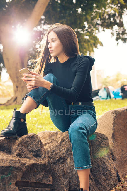 Mujer joven sentada en la roca y sosteniendo el teléfono inteligente en el parque - foto de stock