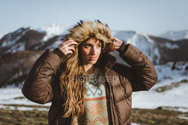 Atractiva mujer que se pone la capucha de chaqueta caliente y mirando hacia otro lado en las montañas nevadas - foto de stock