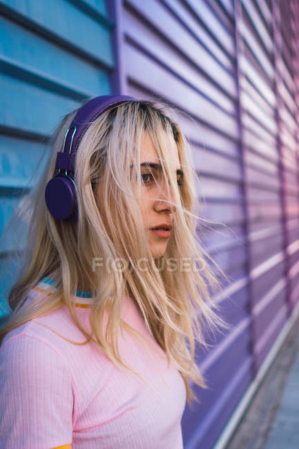 Mujer rubia joven con auriculares morados de pie frente a la pared colorida - foto de stock