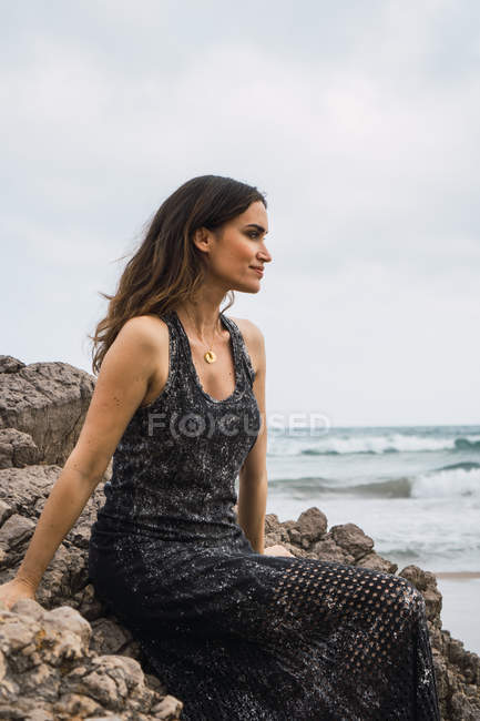 Mujer sonriente pensativa sentada en la roca en el mar - foto de stock