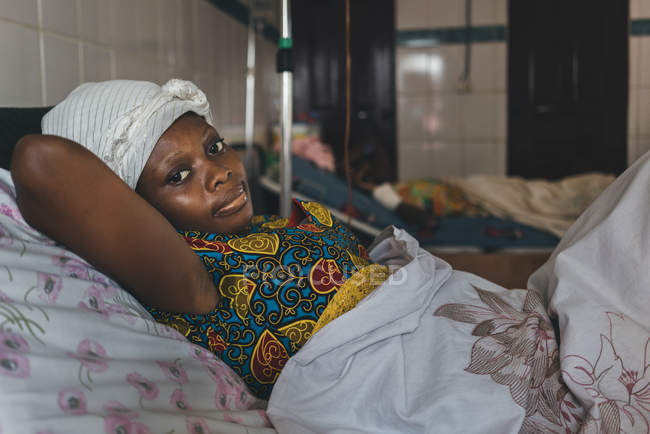 CAMERÚN - ÁFRICA - 5 DE ABRIL DE 2018: Mujer étnica adulta acostada en la cama en el hospital y mirando a la cámara - foto de stock