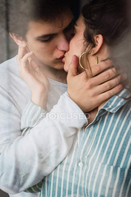 Vista a través de la ventana del joven hombre y la mujer en pijama de pie y besándose - foto de stock