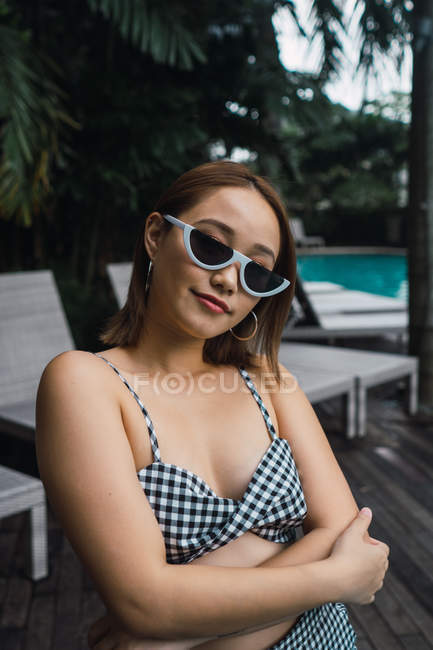 Retrato de mujer asiática joven en gafas de sol de pie en la piscina - foto de stock