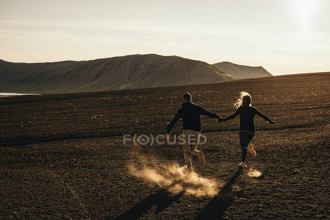 Rückansicht eines Paares, das sich an den Händen hält und abends auf schmutzigem Boden zu den Hügeln rennt. — Stockfoto