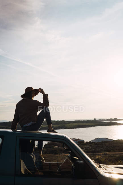 Mulher de chapéu sentado no telhado do carro à beira-mar e olhando para a vista — Fotografia de Stock