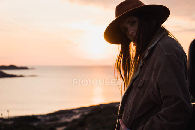 Portrait of dreamy woman in hat and jacket standing at seaside — Stock Photo