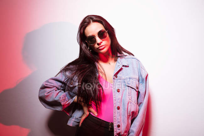 Bella giovane donna vestita di rosa con gli occhiali da sole in piedi al muro bianco e guardando la fotocamera — Foto stock