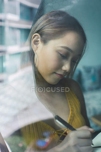 Vista através da janela de mulher asiática bonita de pé e tomar notas — Fotografia de Stock