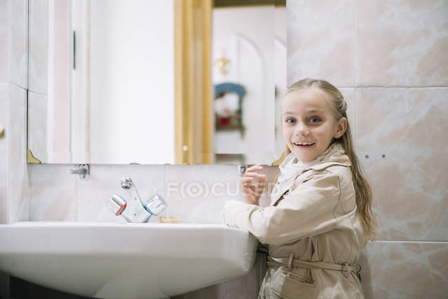 Портрет веселой девушки, стоящей у раковины в пальто и смотрящей в камеру в ванной комнате — стоковое фото