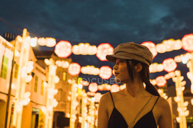 Jeune femme asiatique à la mode regardant loin dans la ville illuminée dans la soirée — Photo de stock