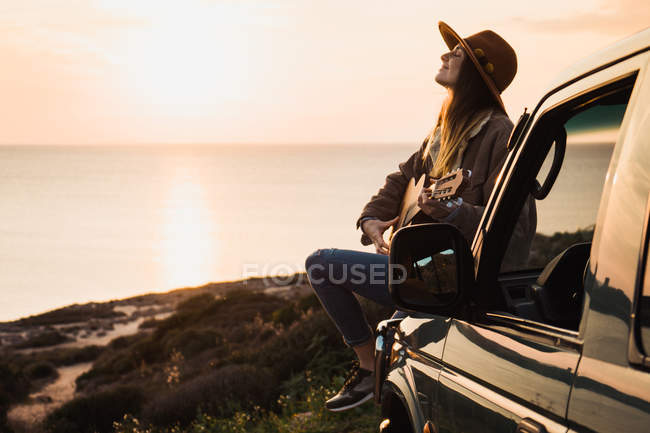 Женщина наслаждается закатом и играет на гитаре, сидя на машине на берегу моря — стоковое фото