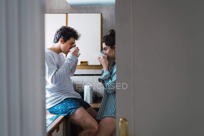 Young couple in pajamas drinking from cups in kitchen — Stock Photo