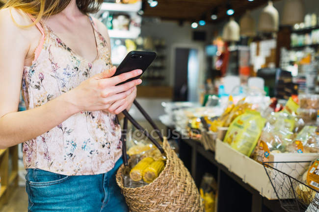Femme avec du maïs dans le panier alimentaire dans l'épicerie en utilisant smartphone — Photo de stock