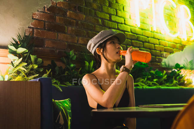 Stilvolle junge Asiatin sitzt im Café und trinkt eine Flasche — Stockfoto