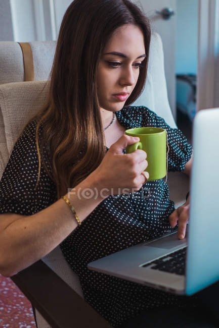 Жінка використовує ноутбук у кріслі і тримає чашку чаю — стокове фото