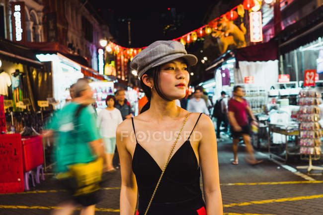 Стильная молодая симпатичная азиатская женщина, идущая по освещенной улице ночью и отводящая взгляд — стоковое фото