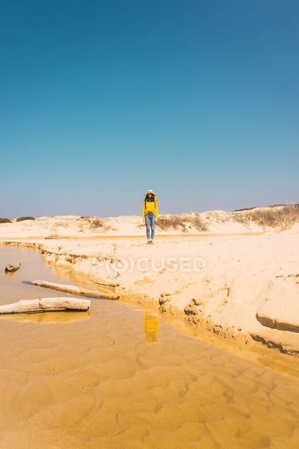 Mulher de chapéu em pé na costa arenosa sob o céu azul — Fotografia de Stock