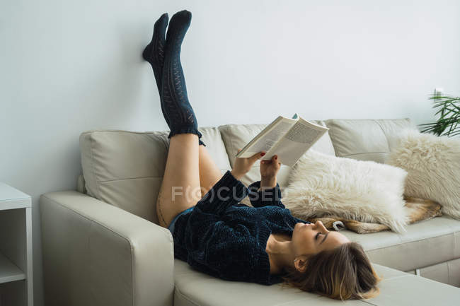 Случайная девушка в свитере и чулках лежит на диване и читает книгу — стоковое фото