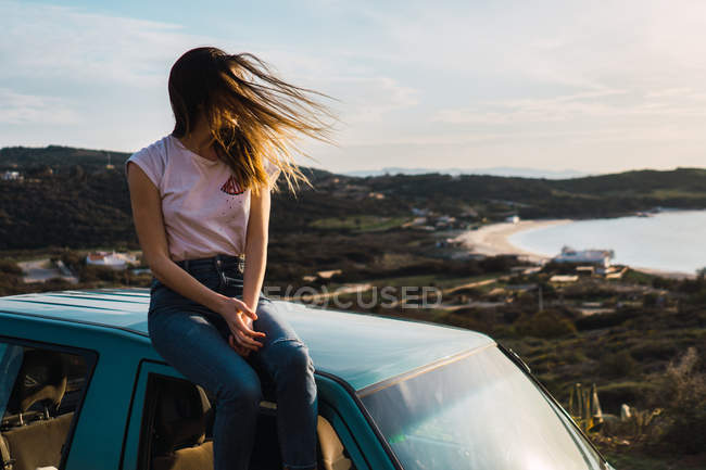 Donna con i capelli ondulati seduta sul tetto dell'auto sulla costa — Foto stock